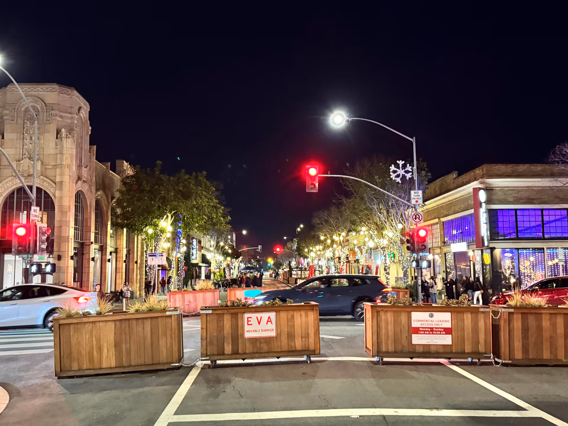 Nighttime downtown street with illuminated storefronts, festive lights, and wooden barriers.