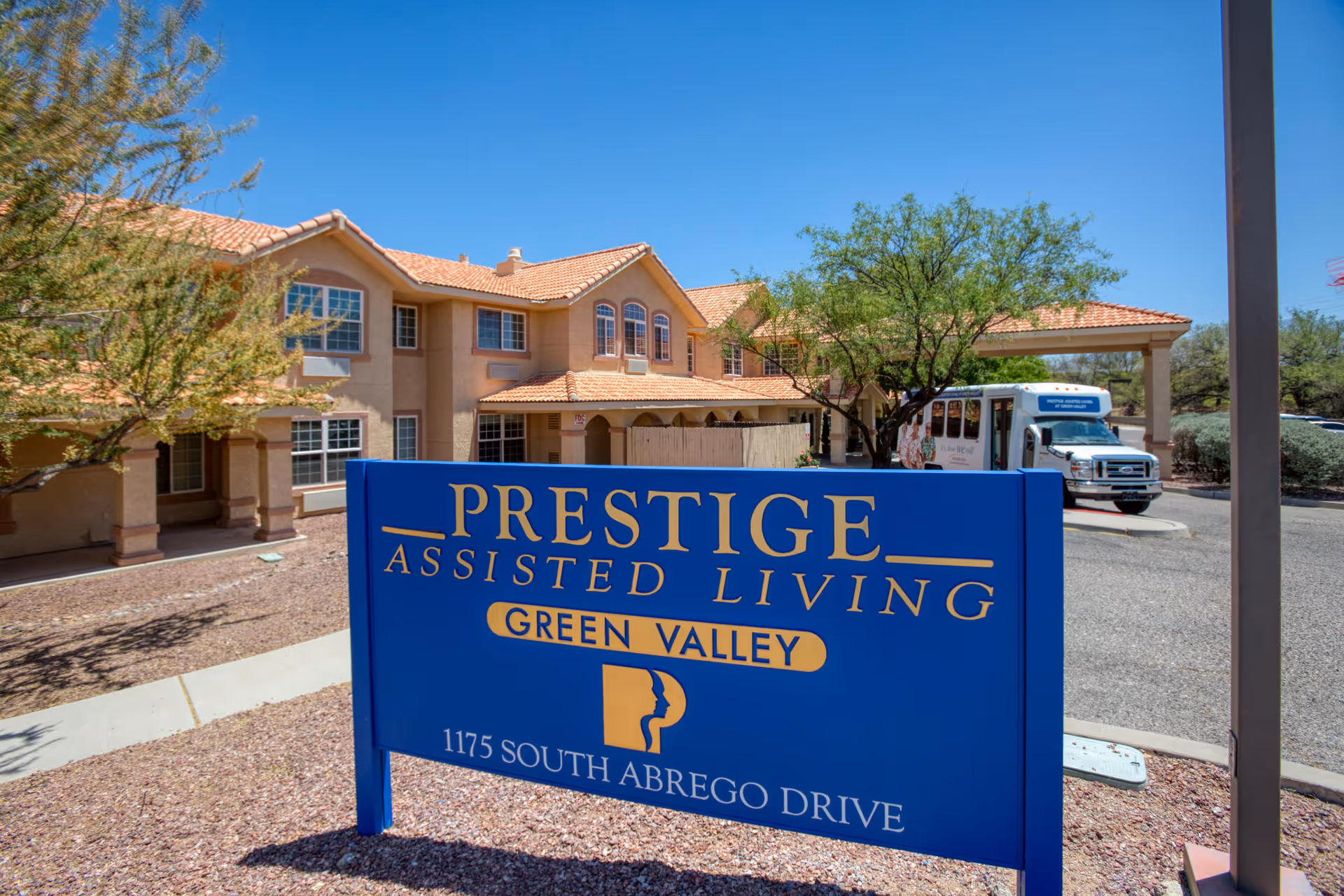 Exterior view of Prestige Assisted Living Green Valley facility with a large blue sign displaying the name and address 1175 South Abrego Drive. The building has a beige stucco exterior with red tile roofing, surrounded by desert landscaping and a clear blue sky. A shuttle bus is parked near the entrance.