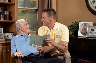Caregiver in a yellow polo sits beside an elderly man, sharing a booklet in living room.