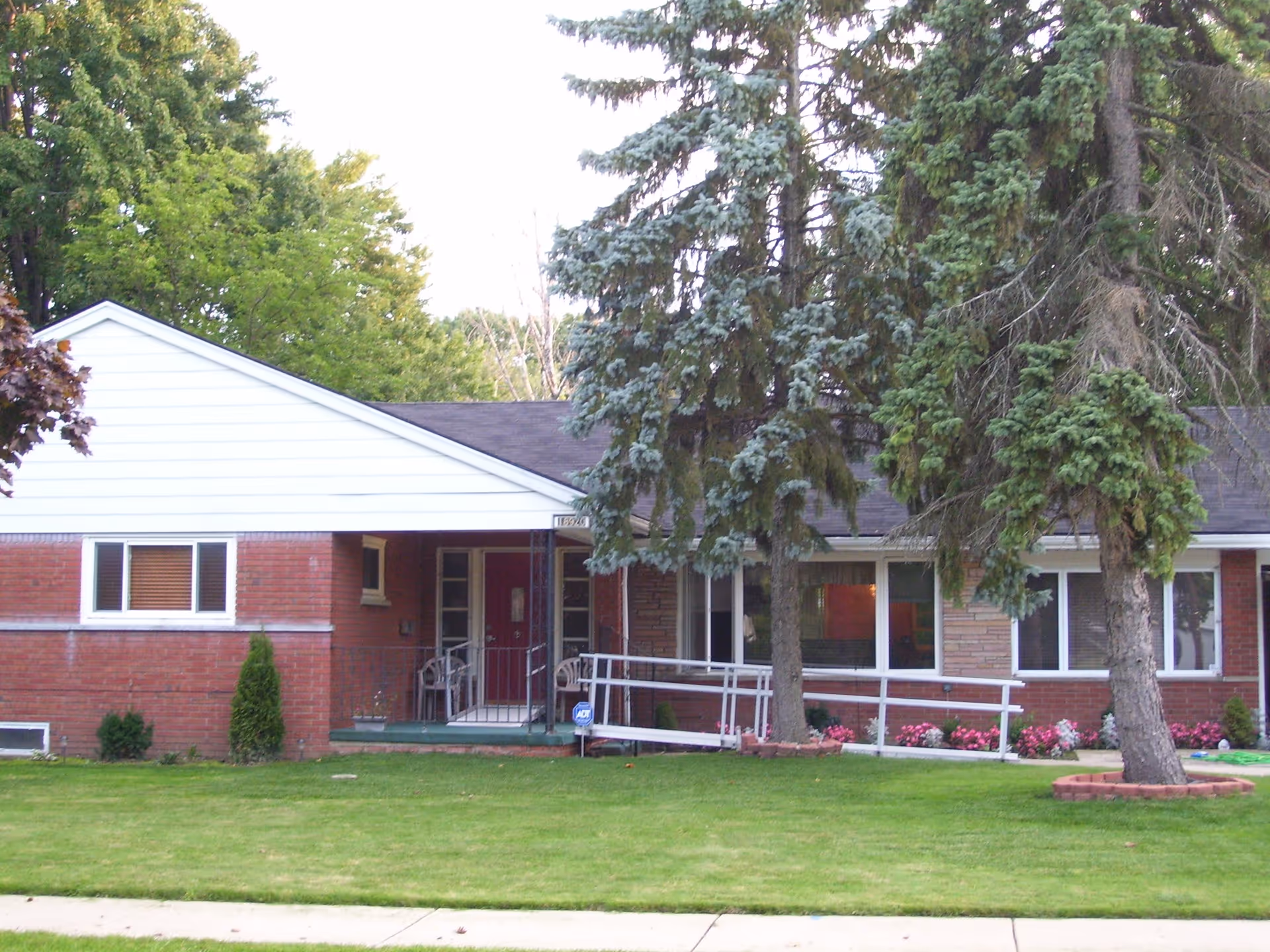 Front view of a single-story brick senior living building with a wheelchair ramp, large pine trees, and a grassy lawn.