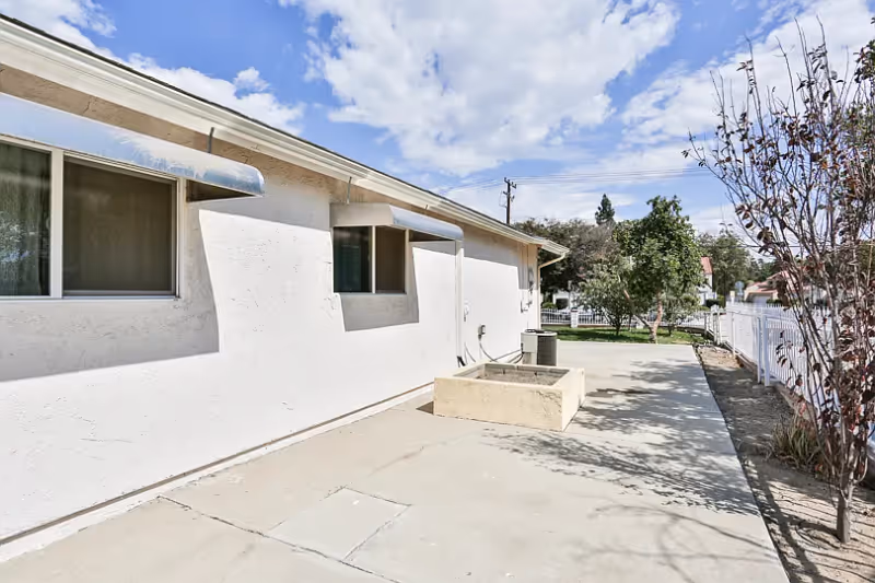 Side exterior of a white stucco single-story building with a concrete patio, a small raised planter, and a white fence under a partly cloudy sky.
