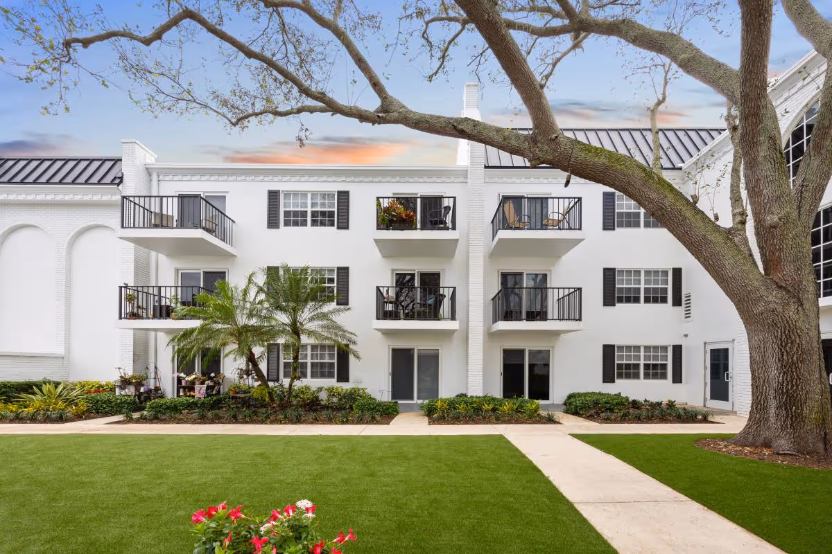 Front exterior of a white three-story senior living building with balconies, landscaped lawn, and a large oak tree in the foreground.