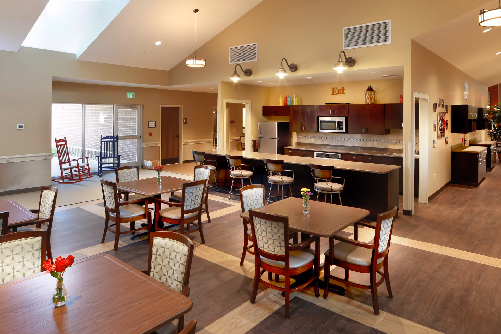 A spacious dining area with several wooden tables and cushioned chairs, each table decorated with a small vase of flowers. In the background, there is a kitchen area with dark wood cabinets, a microwave, refrigerator, and a countertop with bar stools. Two rocking chairs, one red and one blue, are placed near a glass door leading outside. The room has warm lighting and a welcoming atmosphere.