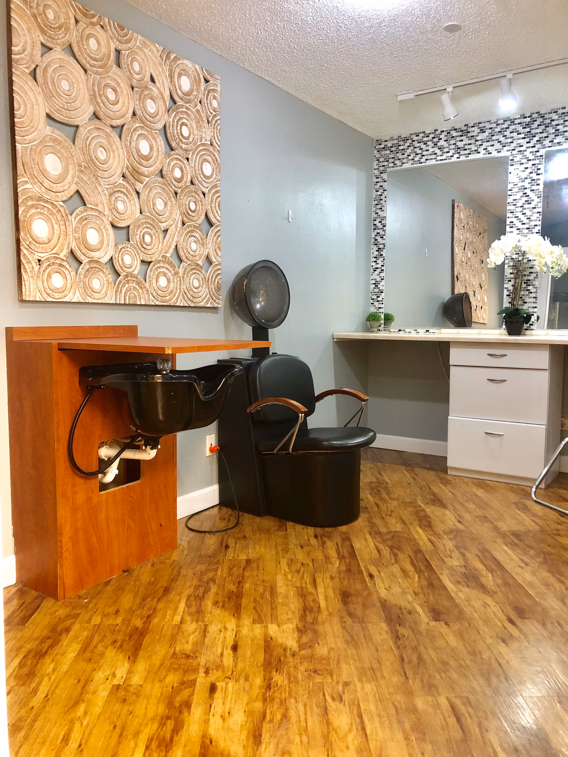 Interior view of a salon area in a senior living facility featuring a black salon chair with wooden armrests next to a black hair washing sink mounted on a wooden cabinet. The wall behind has a large decorative wooden art piece with circular patterns and a large mirror framed with small mosaic tiles. A white countertop with drawers and a potted orchid plant is also visible. The floor is wooden with a warm finish.