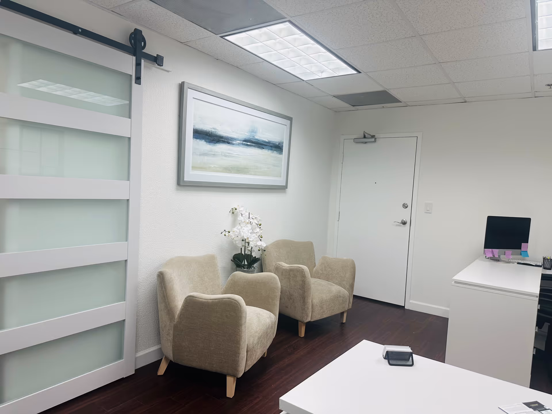 Reception area with beige chairs, white desk, wall art, and frosted glass door.