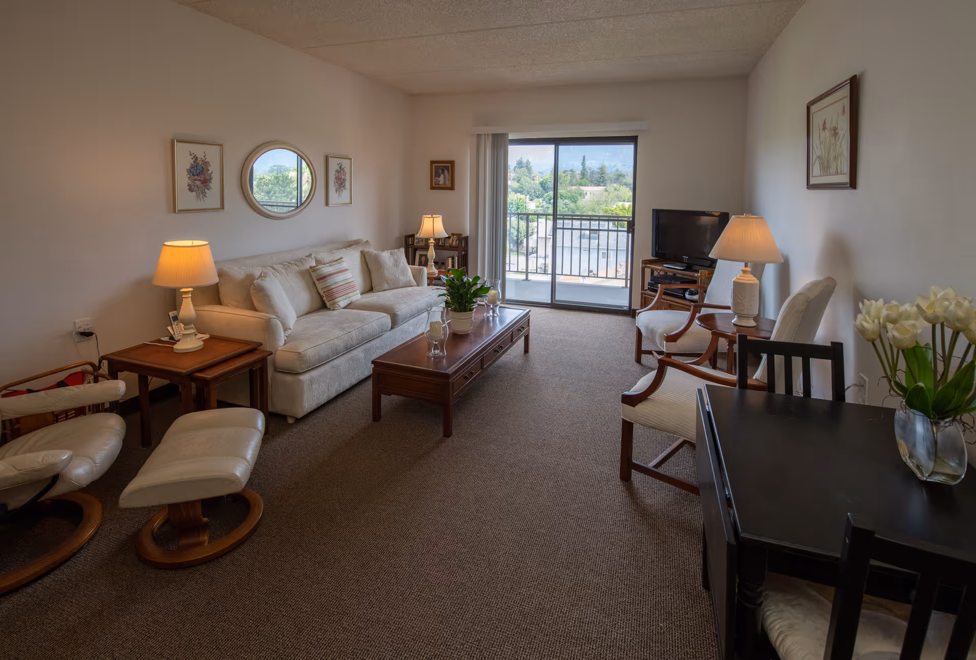 A cozy living room with a beige sofa, a wooden coffee table with a plant and glassware, two armchairs, a TV on a stand, and a sliding glass door leading to a balcony with a view of trees and hills. The room is decorated with framed artwork, table lamps, and a small dining table with chairs and a vase of flowers.