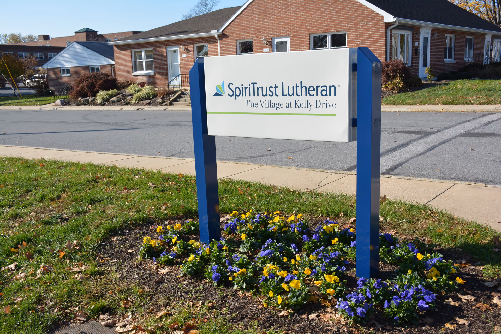 Freestanding 'SpiriTrust Lutheran — The Village at Kelly Drive' sign in a flower bed with a brick building behind it.