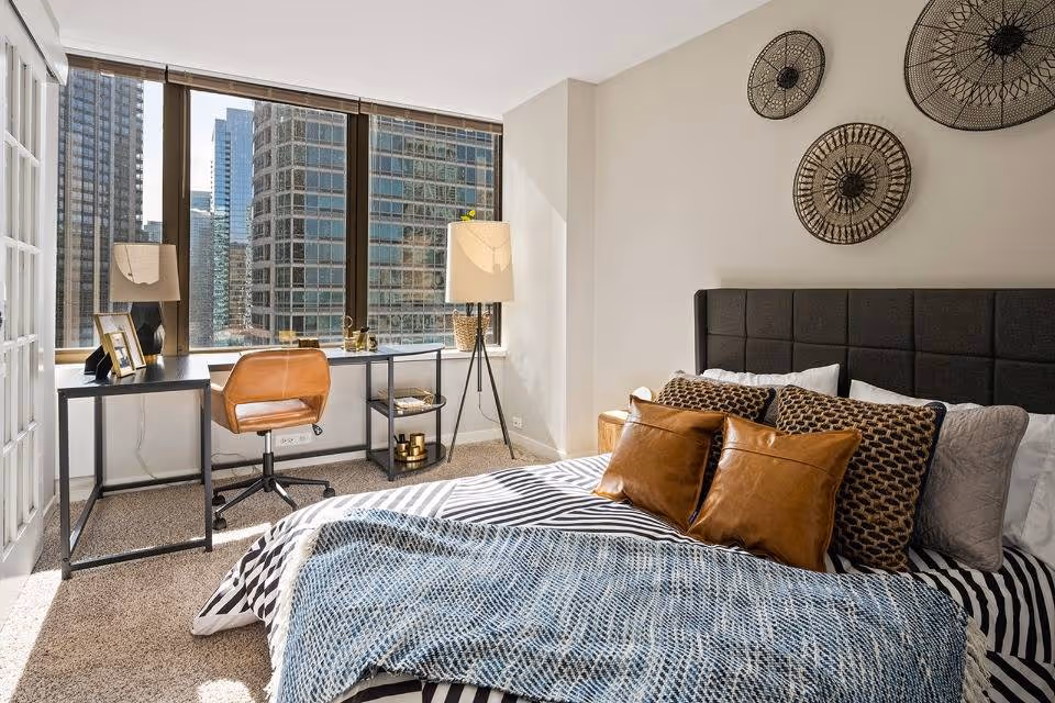 A bright bedroom with a large window showing a cityscape view. The room features a bed with a black upholstered headboard, patterned pillows, and a blue throw blanket. Next to the window is a modern desk with a brown leather chair, a table lamp, and decorative items. The wall above the bed has three circular woven wall hangings.