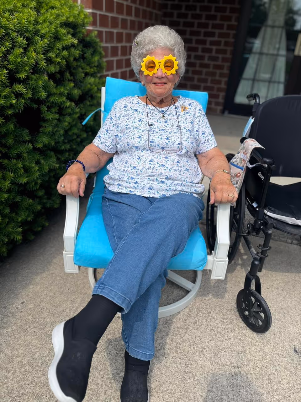 An elderly woman with short gray hair sitting outdoors on a blue cushioned chair. She is wearing a white floral shirt, blue jeans, black socks, and white shoes. She has on playful yellow sunglasses shaped like sunflowers and is smiling. There is a wheelchair next to her and a green bush and brick wall in the background.