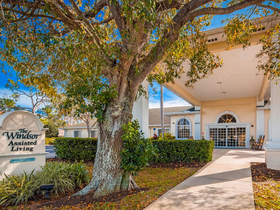 Front entrance of The Windsor Assisted Living with a covered porte-cochere, a large tree, walkway, and signage.