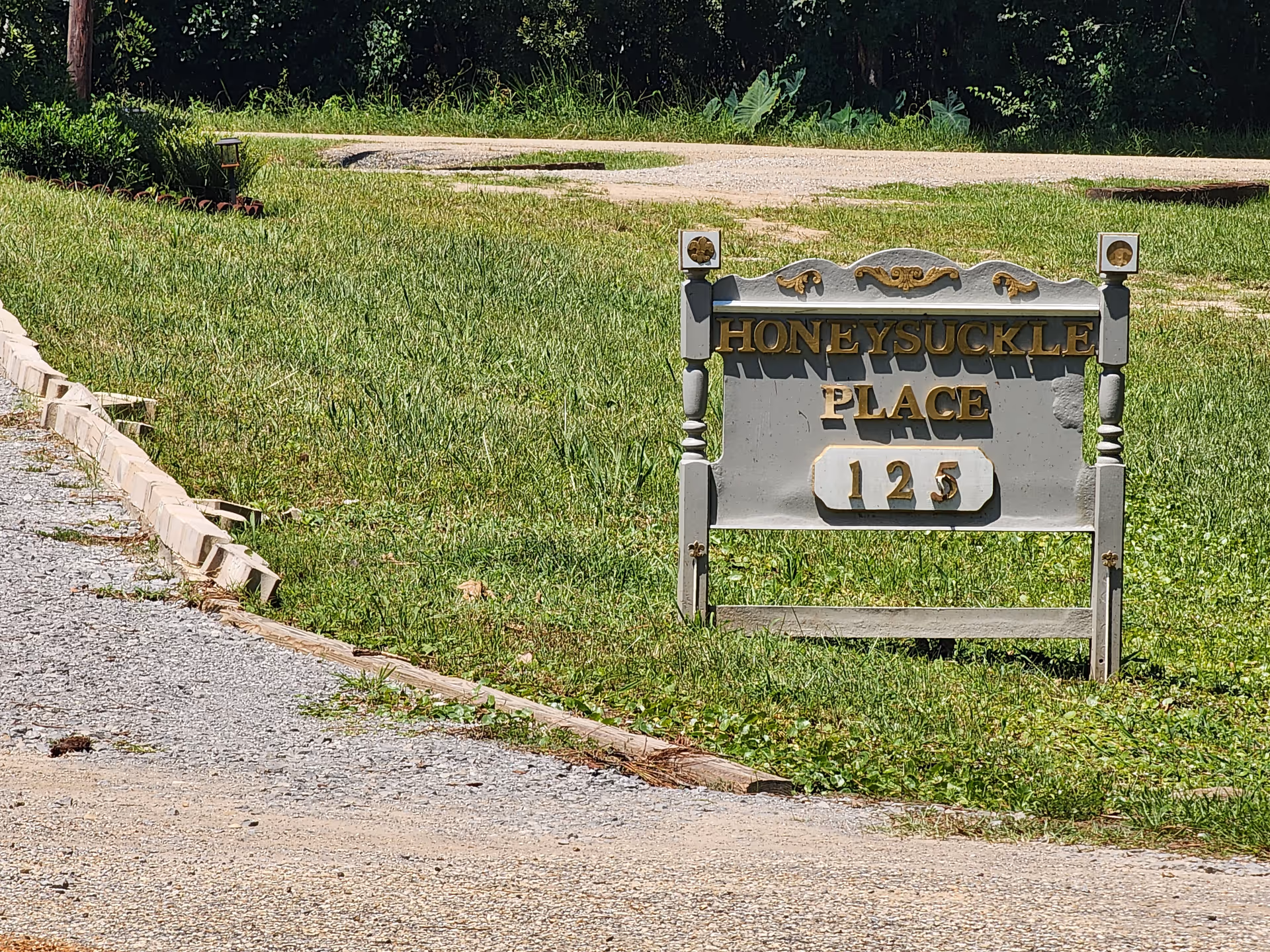 A weathered Honeysuckle Place sign sits on a grassy lawn by a gravel path, 125.