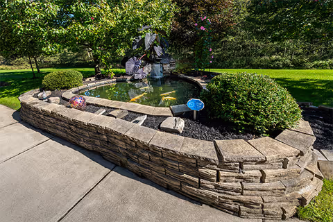 A landscaped outdoor area featuring a stone-bordered pond with a small water fountain in the center, surrounded by green bushes, trees, and a well-maintained lawn under bright sunlight.