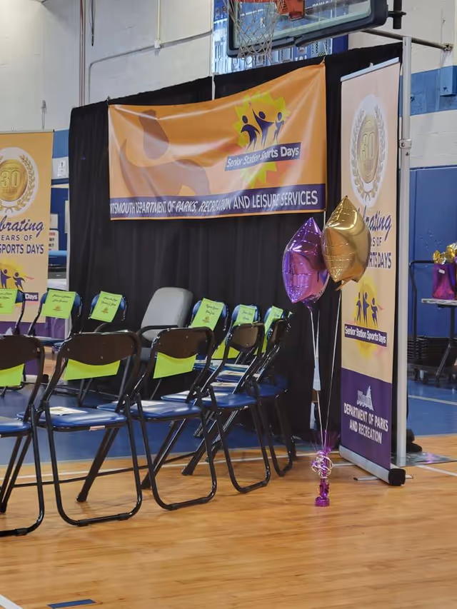 Indoor gym setup with folding chairs, banners, and balloons for senior sports day event.