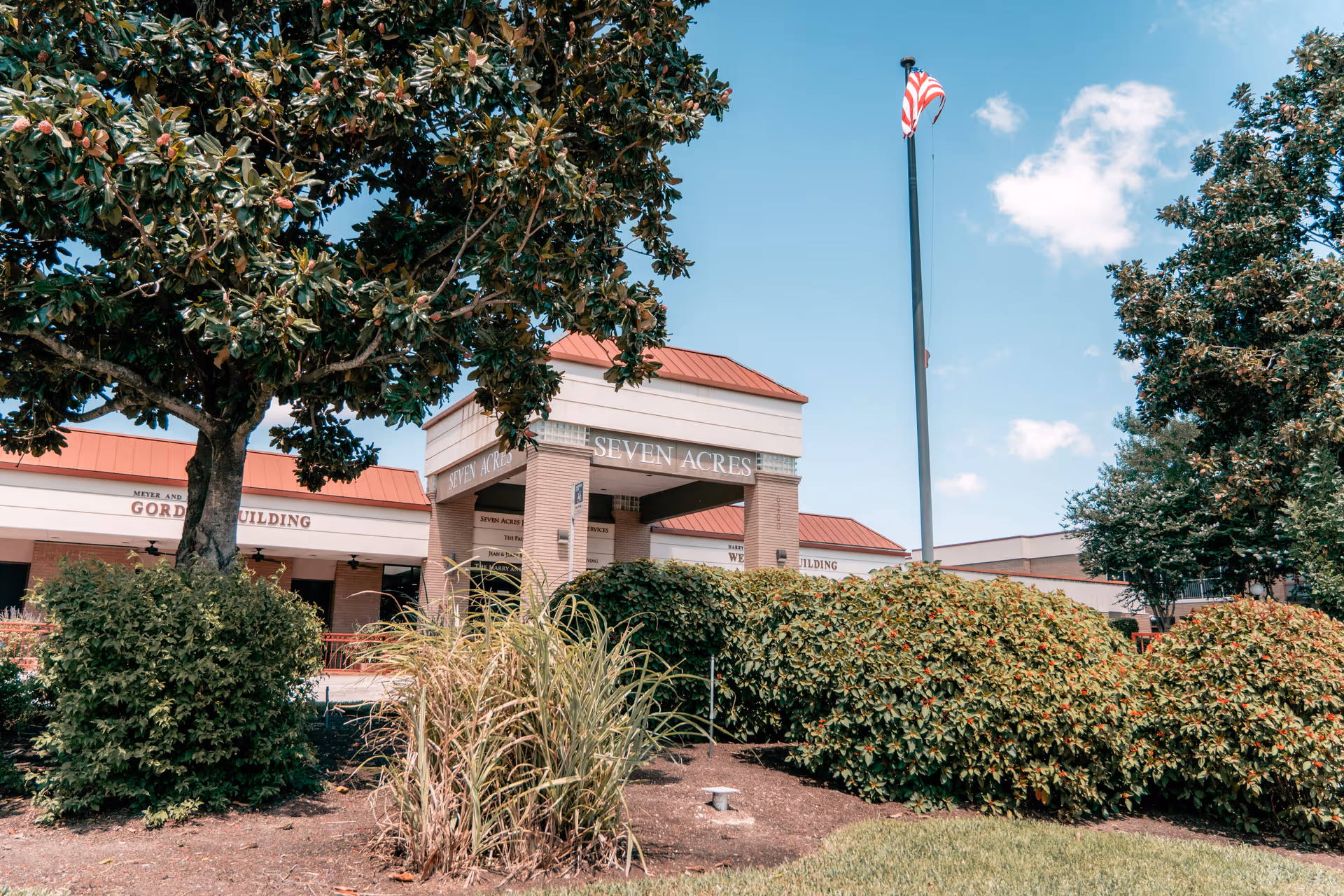 Front entrance of the Seven Acres senior care building with trees, shrubs, and an American flag in front.