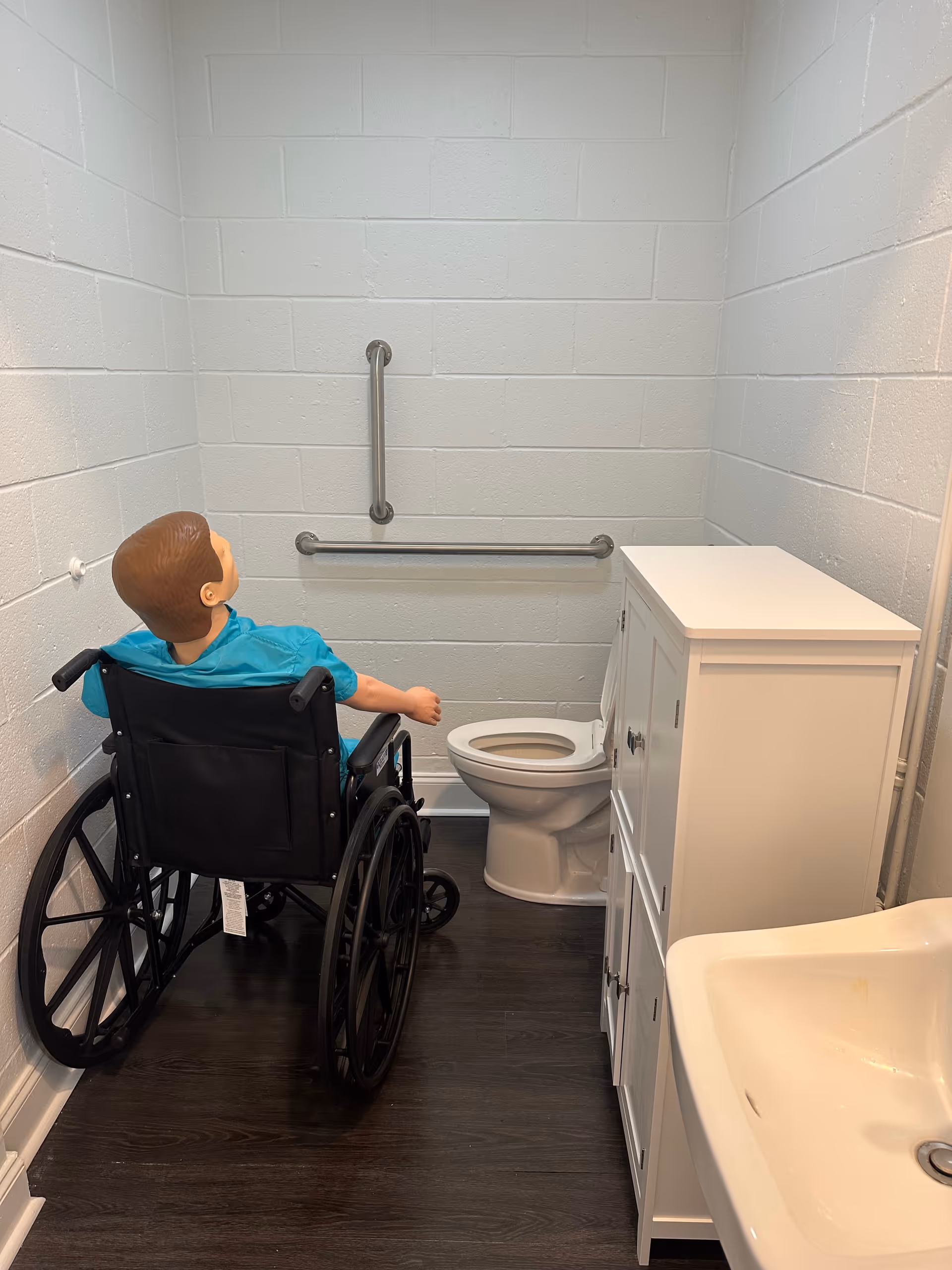 Wheelchair user sits near a toilet in a small bathroom with grab bars and sink.