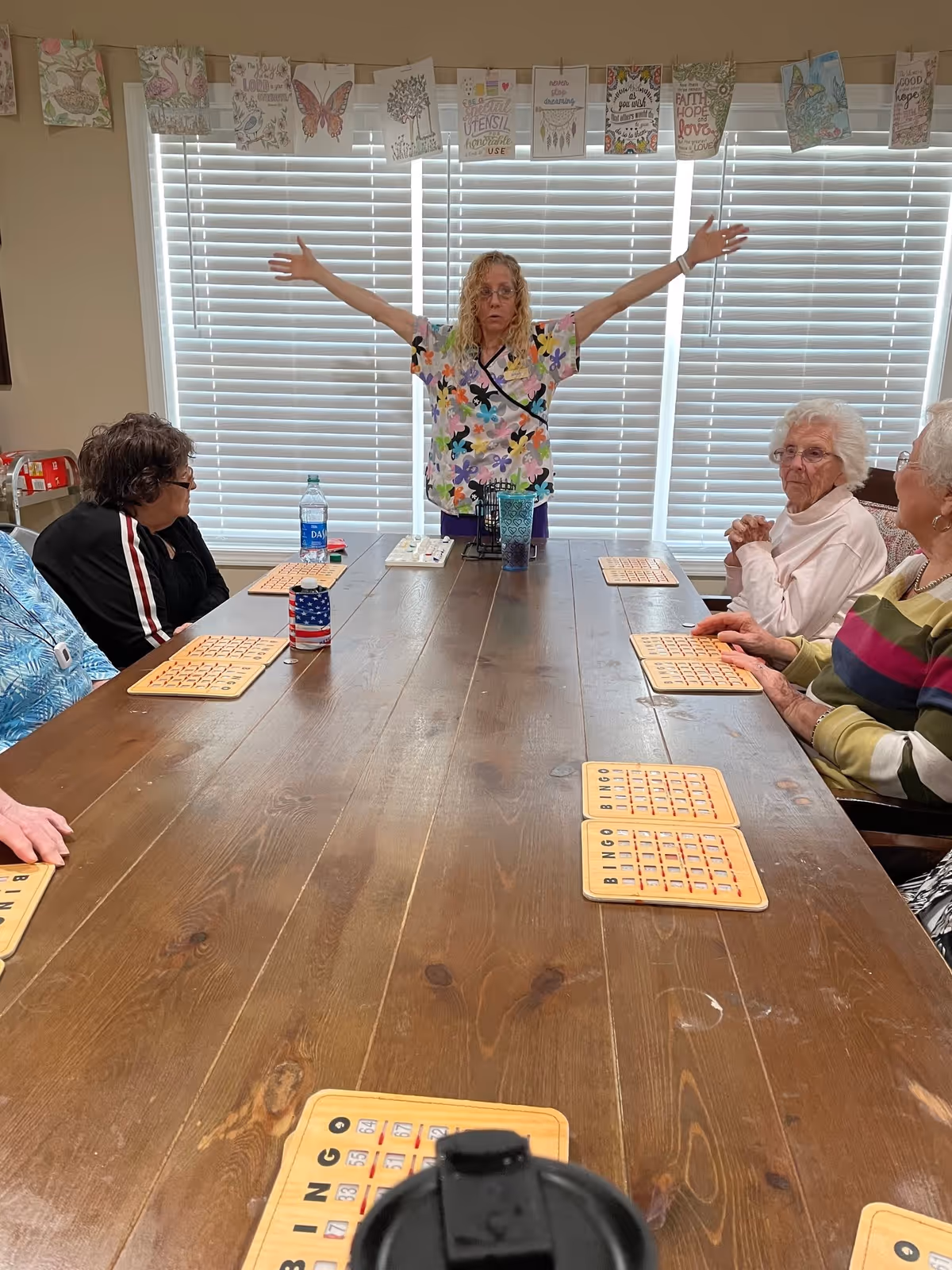 A group of elderly women sitting around a wooden table playing bingo with bingo cards in front of them. A woman standing at the head of the table with arms outstretched appears to be calling out bingo numbers. The room has closed white blinds and decorative cards hanging above the window.