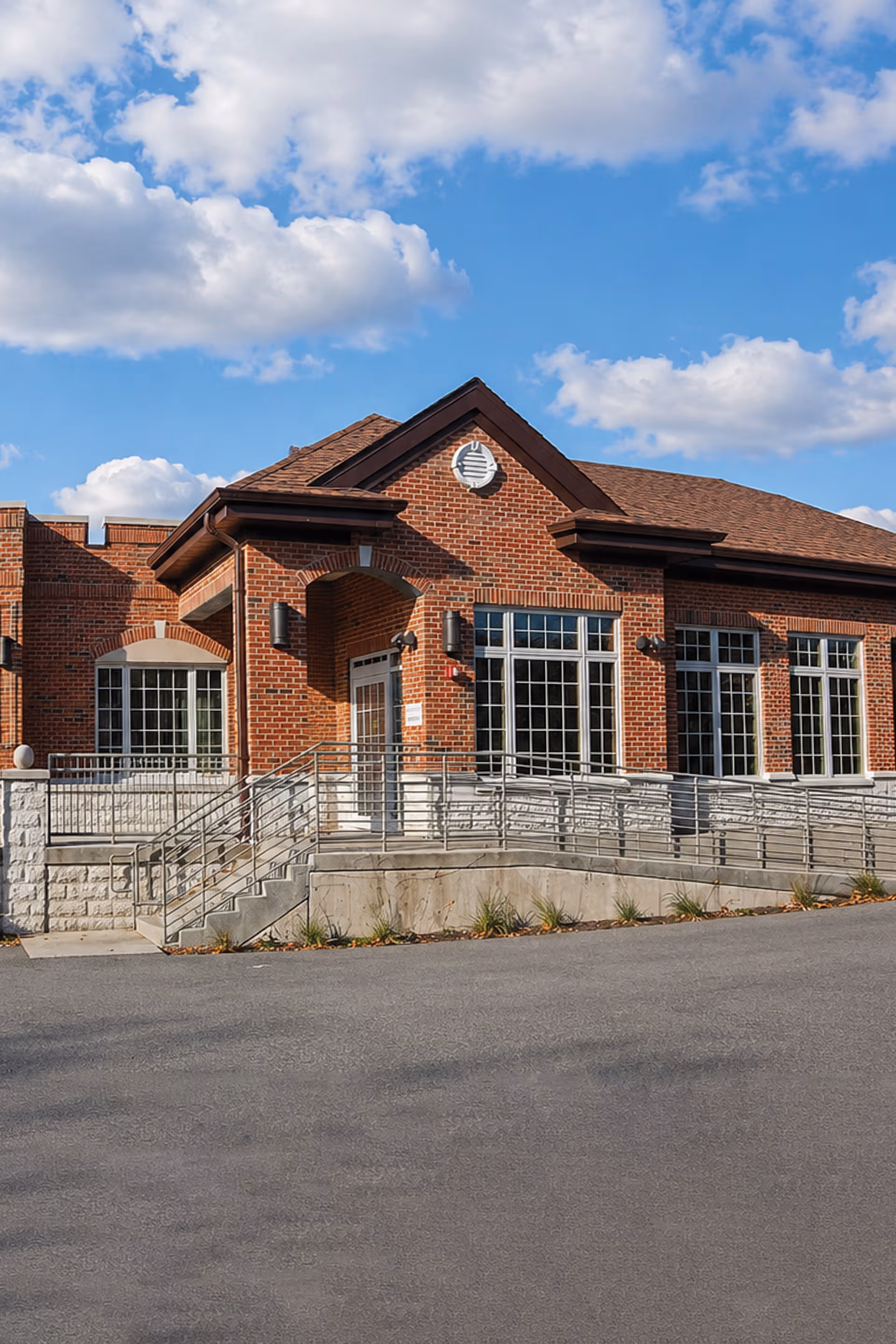 Red brick senior living facility entrance with ramp and large windows under a blue sky.