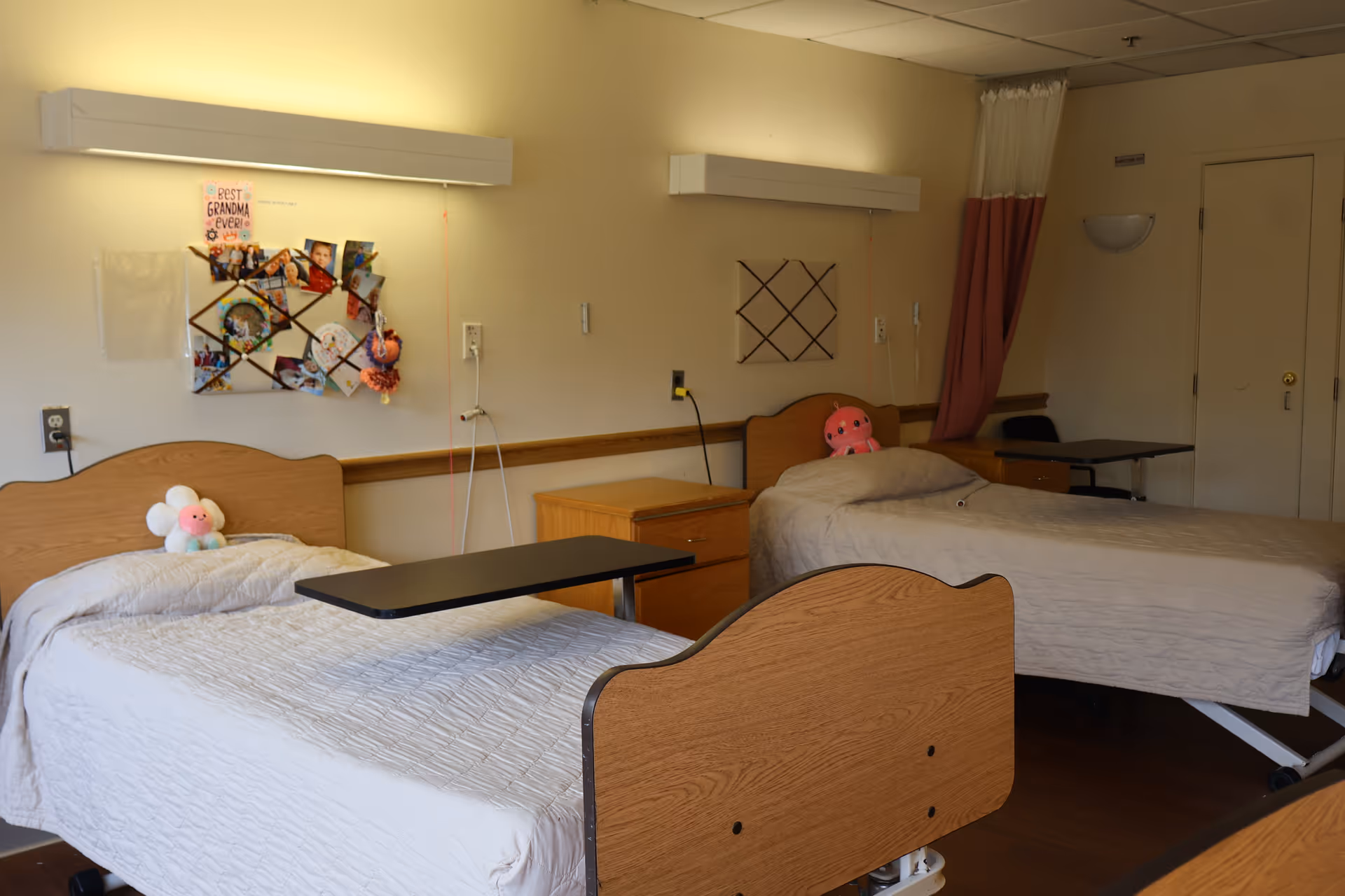 A room with two hospital-style beds, each with a small stuffed toy on the pillow. There are bedside tables between and beside the beds, and a bulletin board with photos and notes on the wall above one bed. The room has a curtain partially drawn near the door and soft lighting.