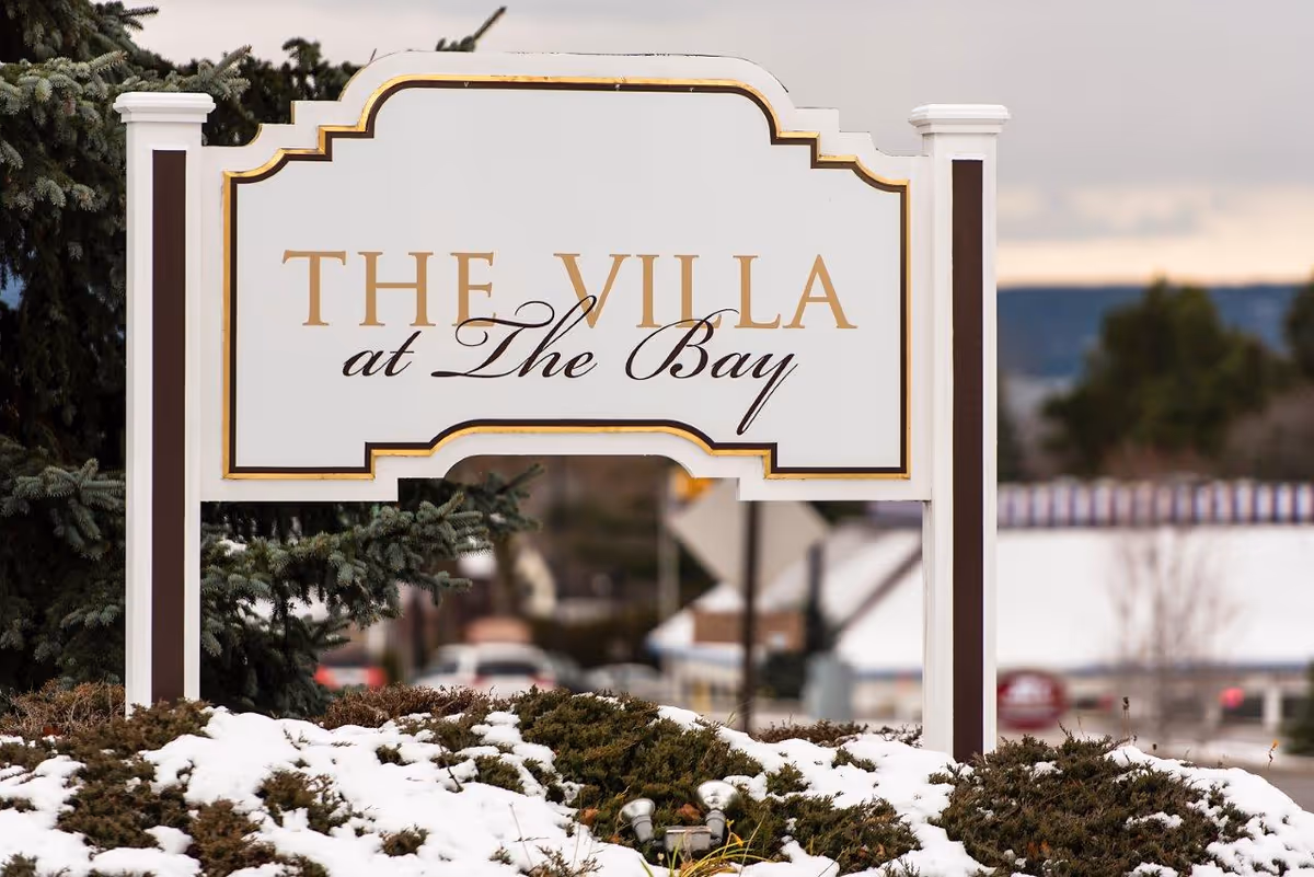 White and gold sign reading "The Villa at The Bay" mounted on posts in a snowy landscaped area.
