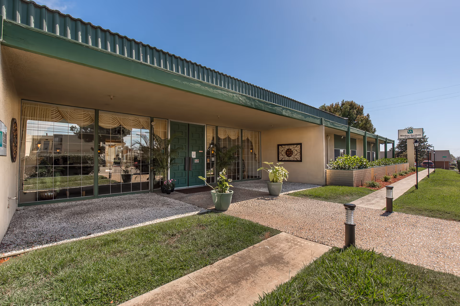 Front entrance of a single-story convalescent center with green trim, potted plants, large windows, and a paved walkway.
