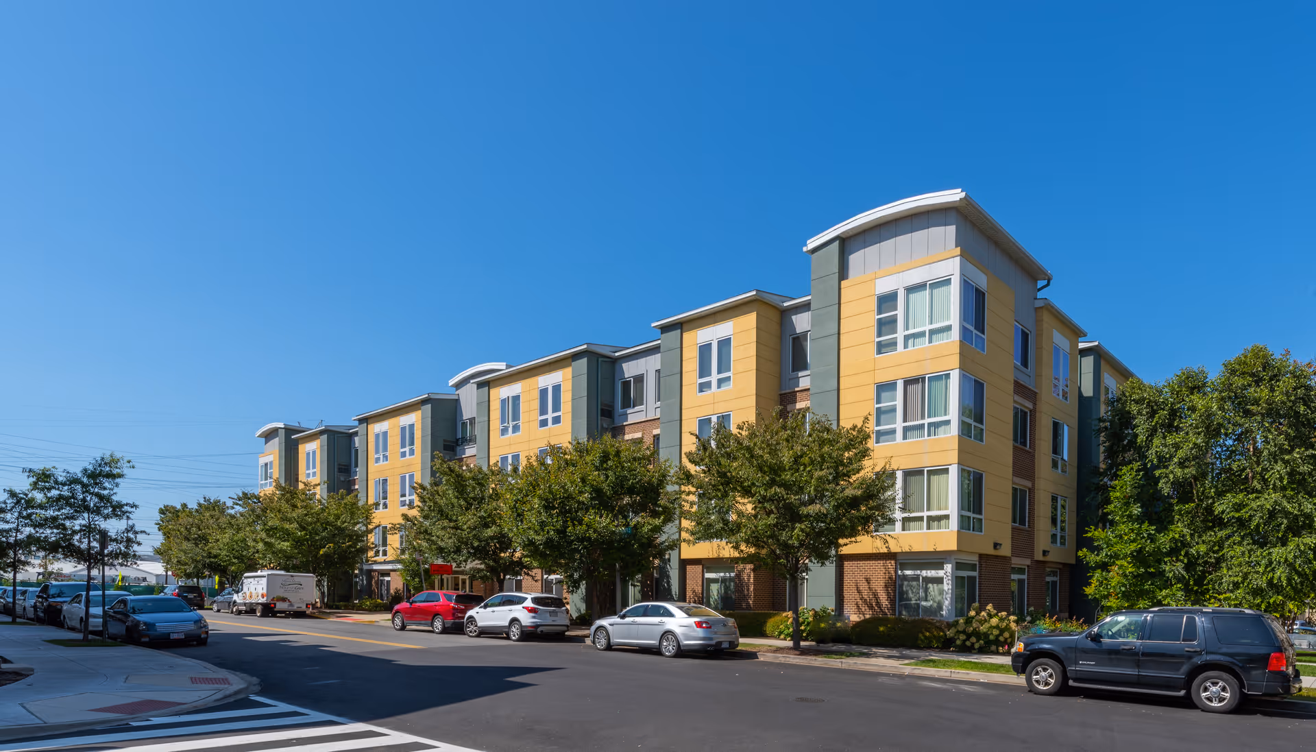 Colorful multi-story senior living building with trees, parked cars, and a clear blue sky.