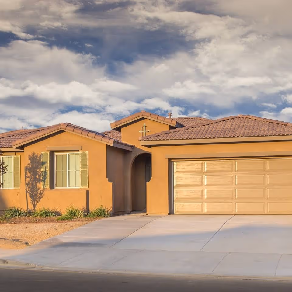 Front exterior of a single-story stucco house with a tiled roof, two-car garage, and driveway under a cloudy sky.