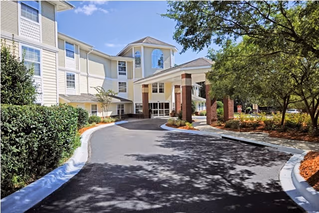 Exterior view of Allegro Senior Living in Tallahassee, FL, showing a multi-story building with beige siding and white trim. The entrance features a covered driveway supported by brick columns. Trees and landscaping surround the driveway and building under a clear blue sky.