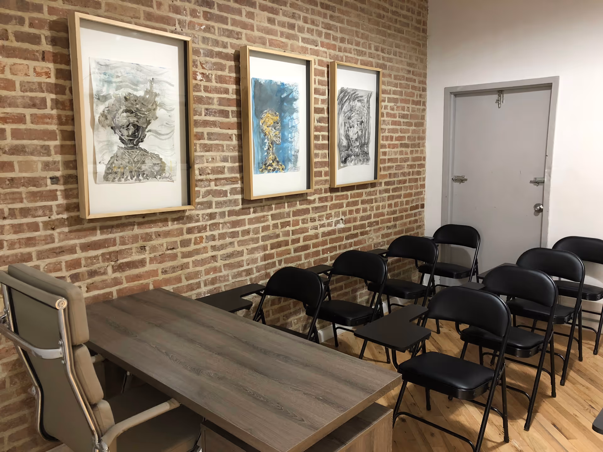 Brick-walled interior conference room with a wooden desk, framed art, and rows of black chairs.