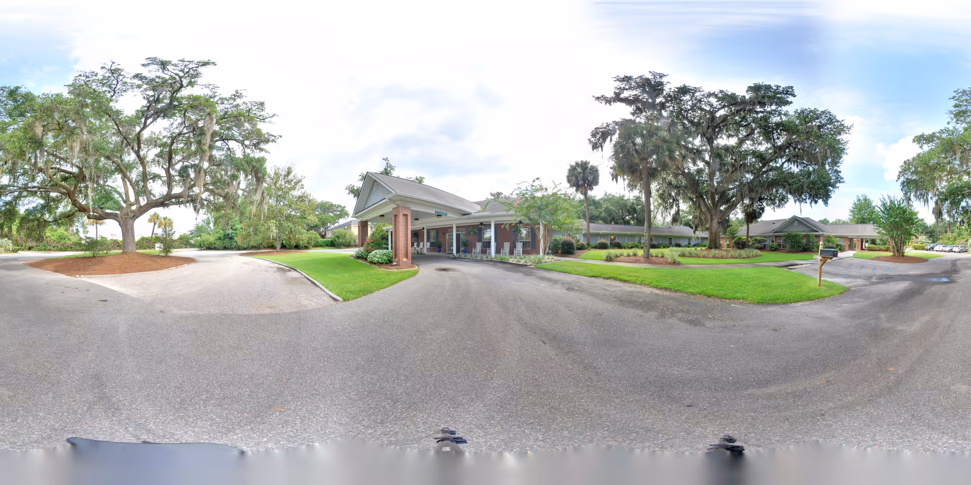 Wide panoramic view of the front exterior of Oaks at Beaufort senior living facility, showing a driveway with a covered entrance, surrounded by well-maintained green lawns, large trees, and a partly cloudy sky.