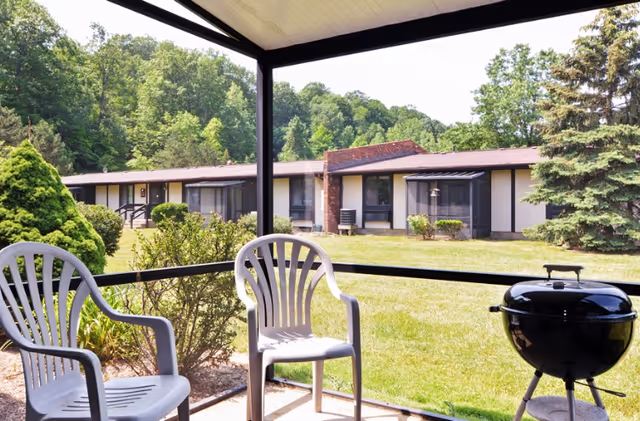 View from a screened porch with white plastic chairs, a charcoal grill, and a grassy courtyard.