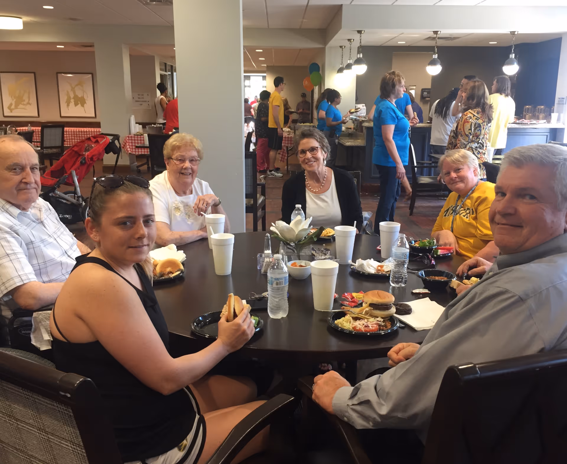 Several people sit around a round table eating burgers and drinks in a communal dining area while others mingle in the background.
