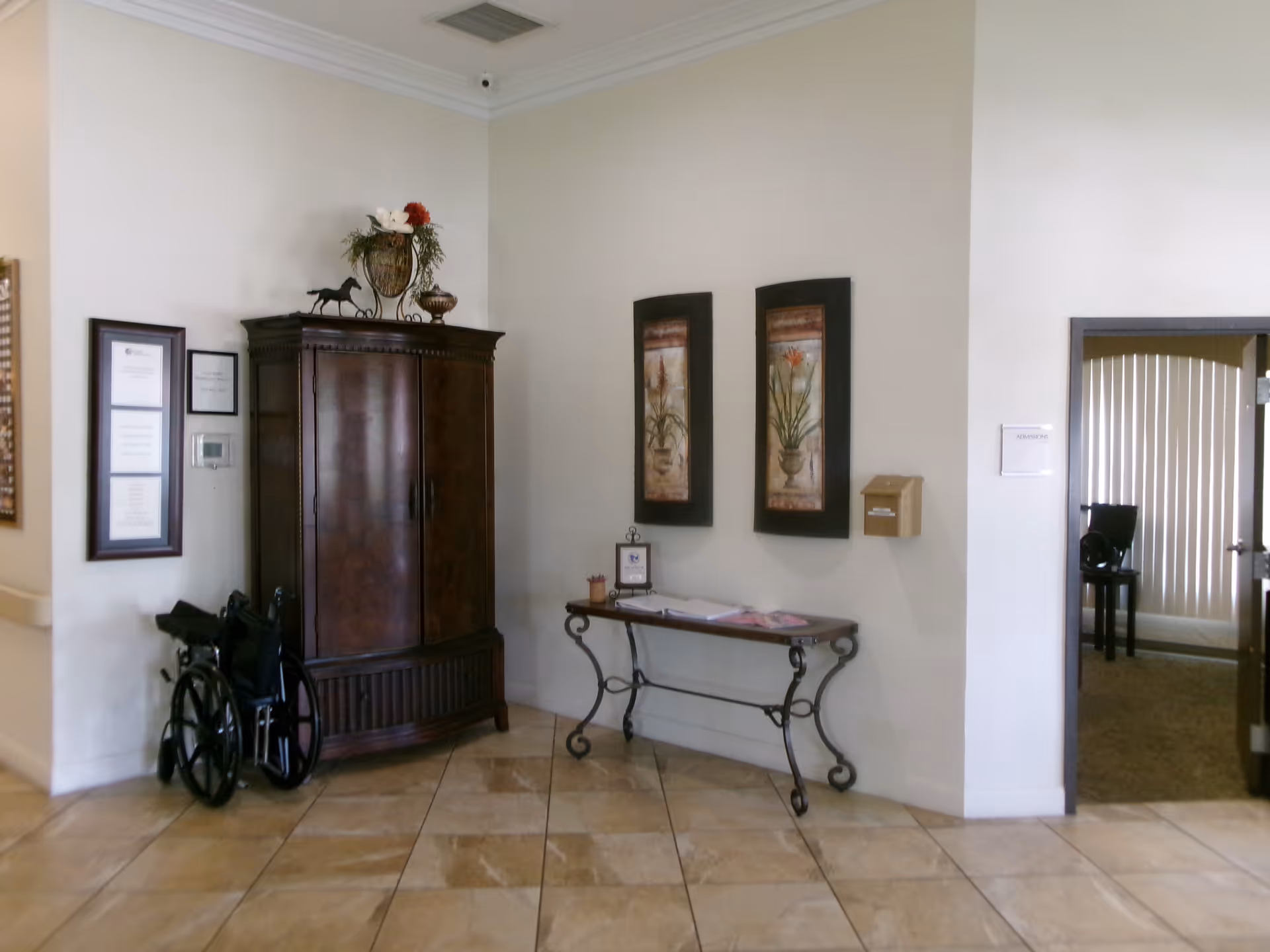 Interior corner of a facility with a dark wooden cabinet, a wheelchair beside it, a decorative table with papers on it, two framed floral paintings on the wall, and a doorway leading to another room with vertical blinds.