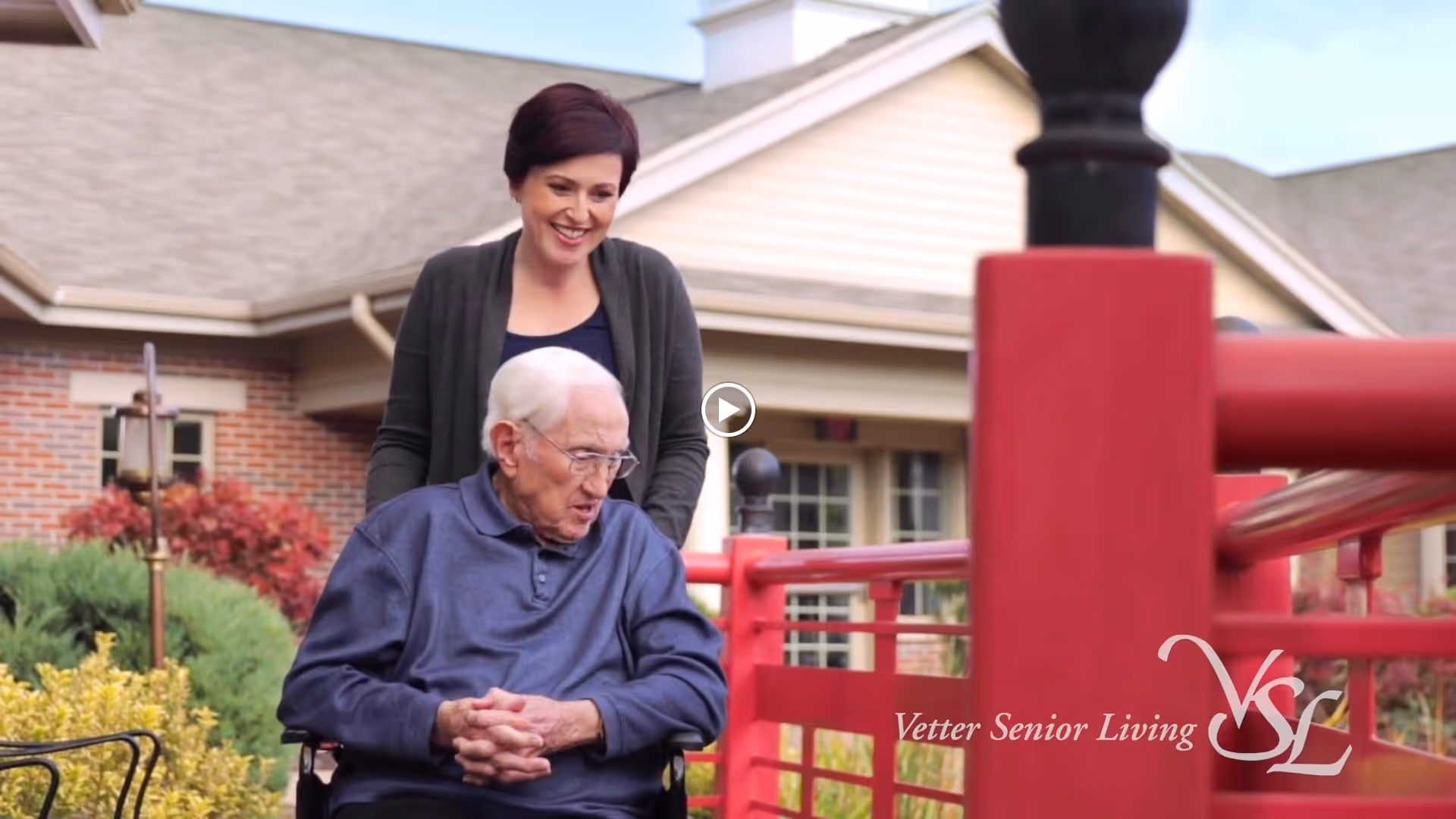 An elderly man in a wheelchair is outdoors near a red railing, accompanied by a smiling woman standing behind him. They are in front of a building with brick and beige siding, surrounded by greenery and shrubs.
