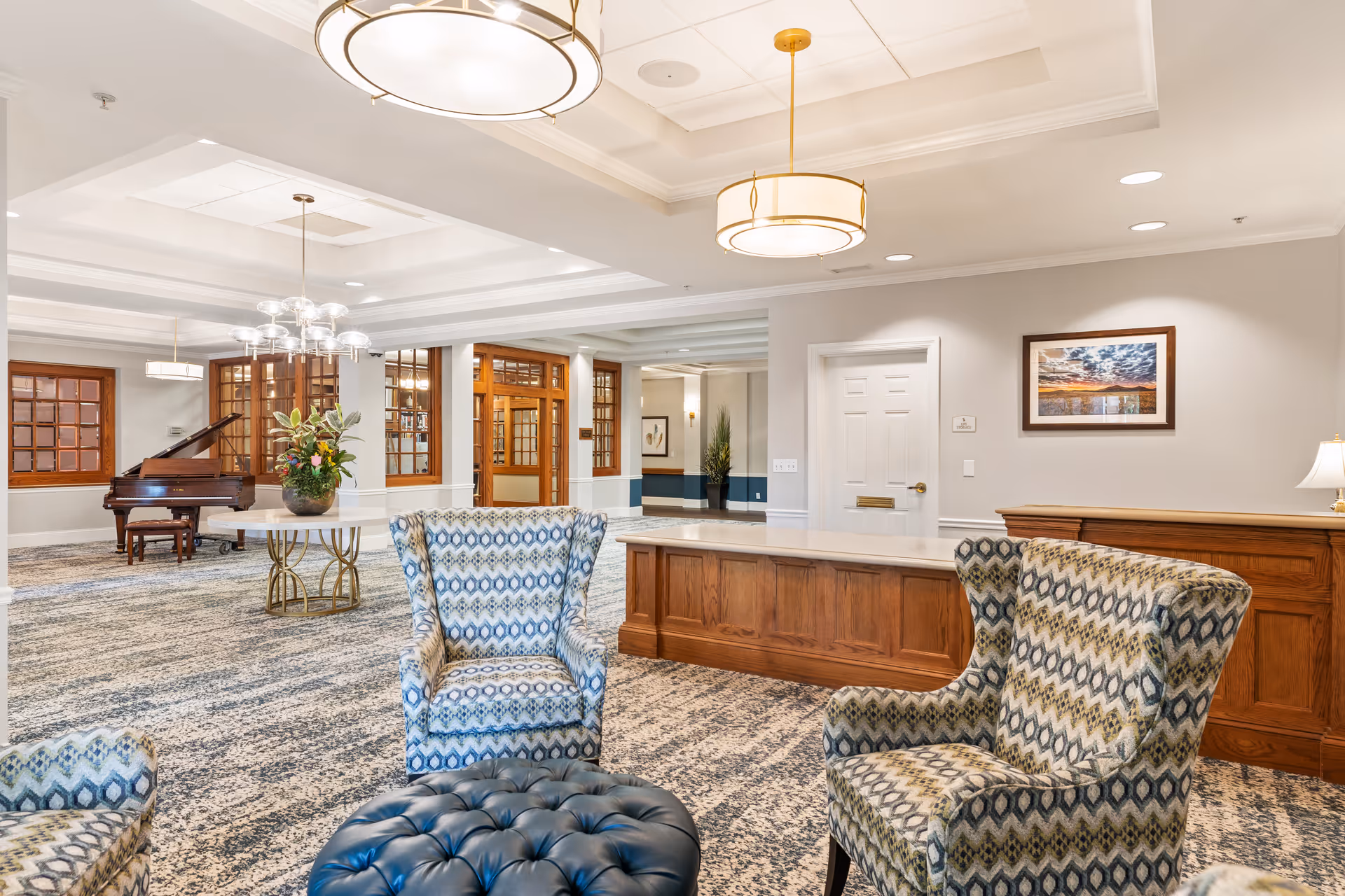 A spacious and well-lit common area in a senior living facility featuring patterned armchairs around a tufted ottoman, a wooden reception desk, a round table with a floral arrangement, and a grand piano in the background. The room has large windows with wooden frames, modern ceiling lights, and a framed landscape photograph on the wall.
