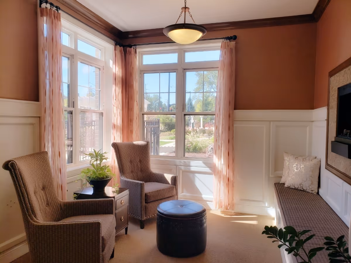 Sunlit seating nook with two armchairs, a bench, plant, and fireplace along paneled walls.