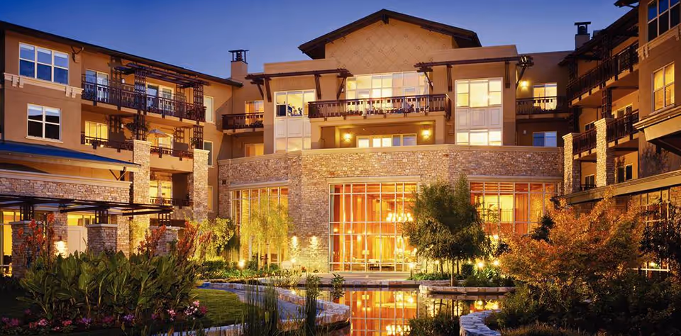 Illuminated multi-story senior living building with balconies, landscaped grounds and a reflecting pond at dusk.