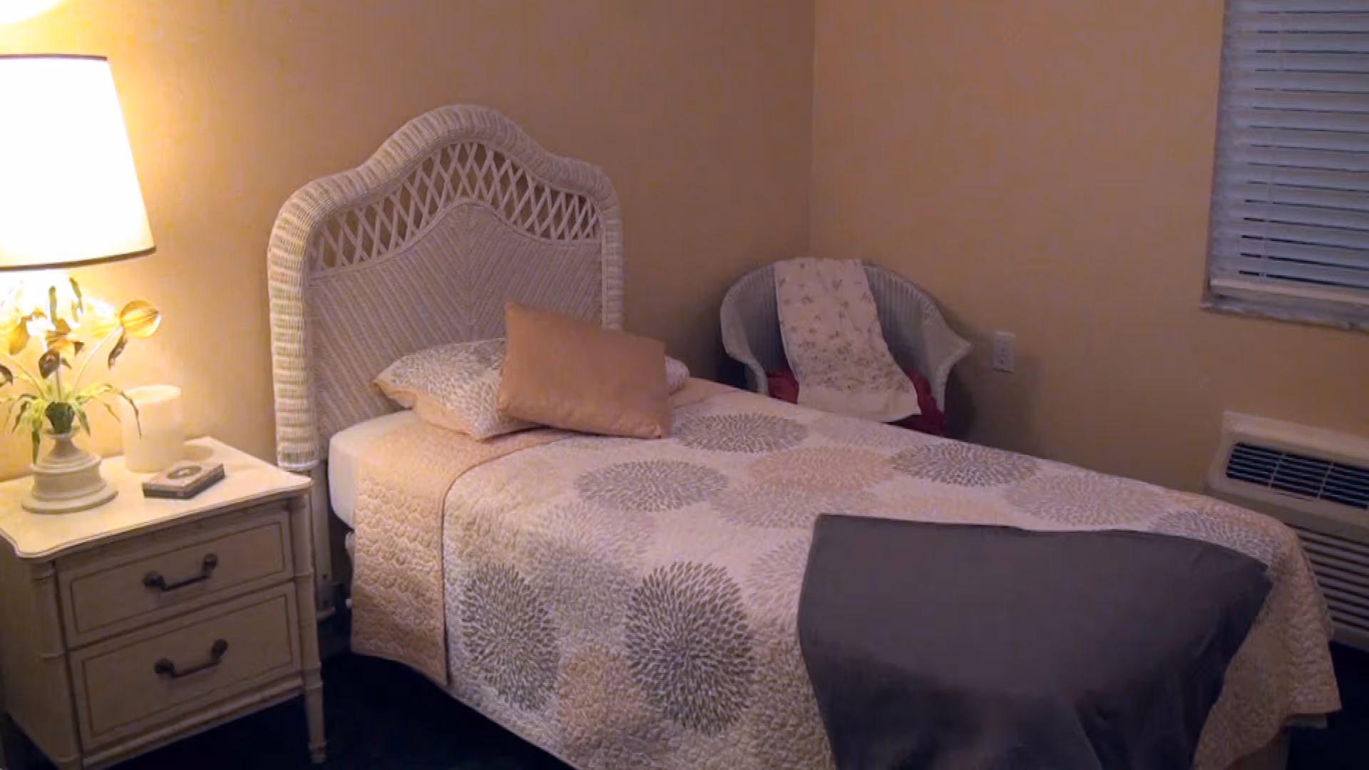 A softly lit bedroom with a single bed topped with a patterned quilt, a nightstand with a lamp and flowers, and a wicker chair.
