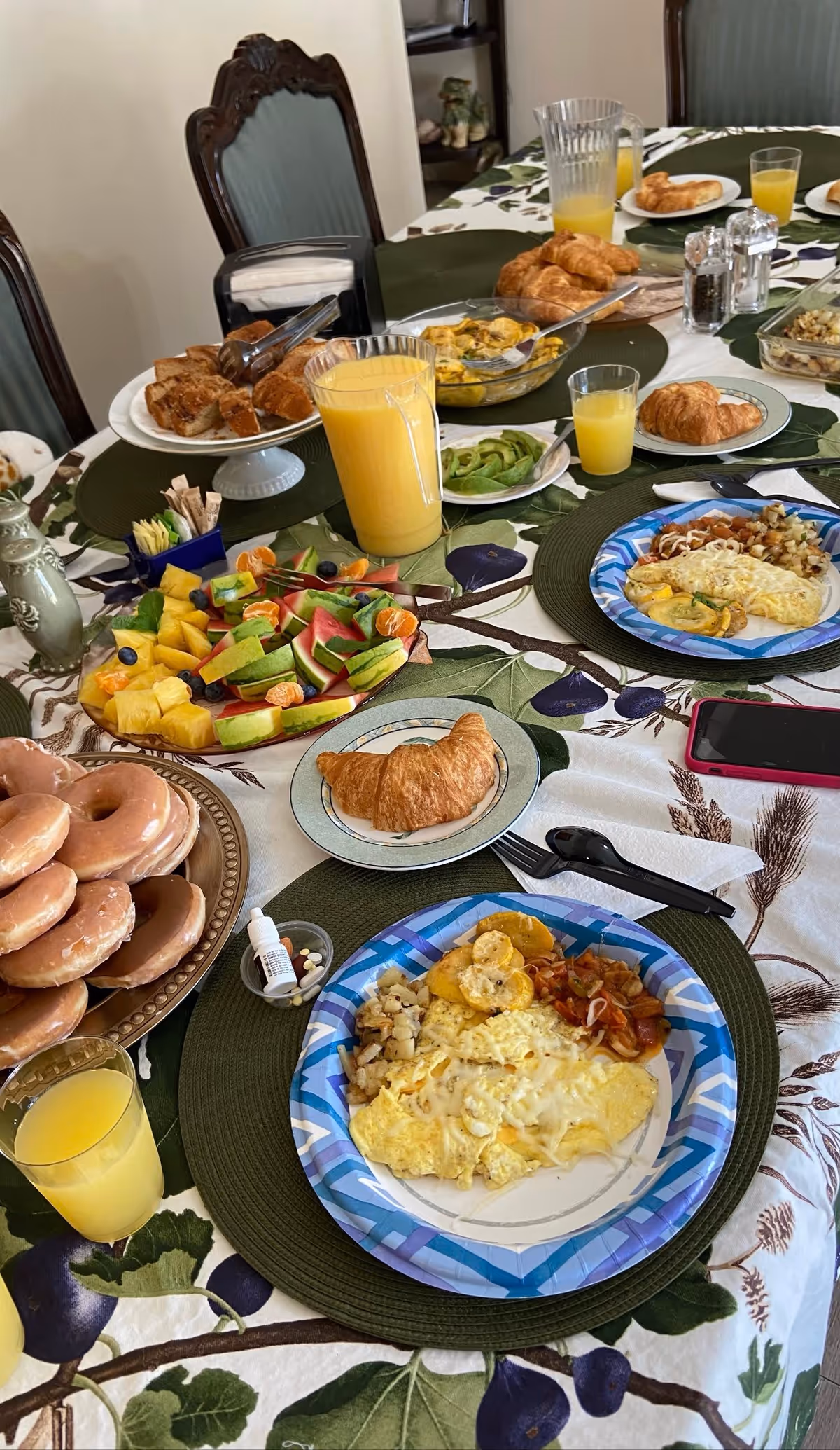 A dining table set for breakfast with plates of omelets, croissants, a platter of assorted fresh fruit, a stack of glazed donuts, glasses and a pitcher of orange juice, and various condiments. The table has a floral tablecloth and green placemats, with wooden chairs around it.