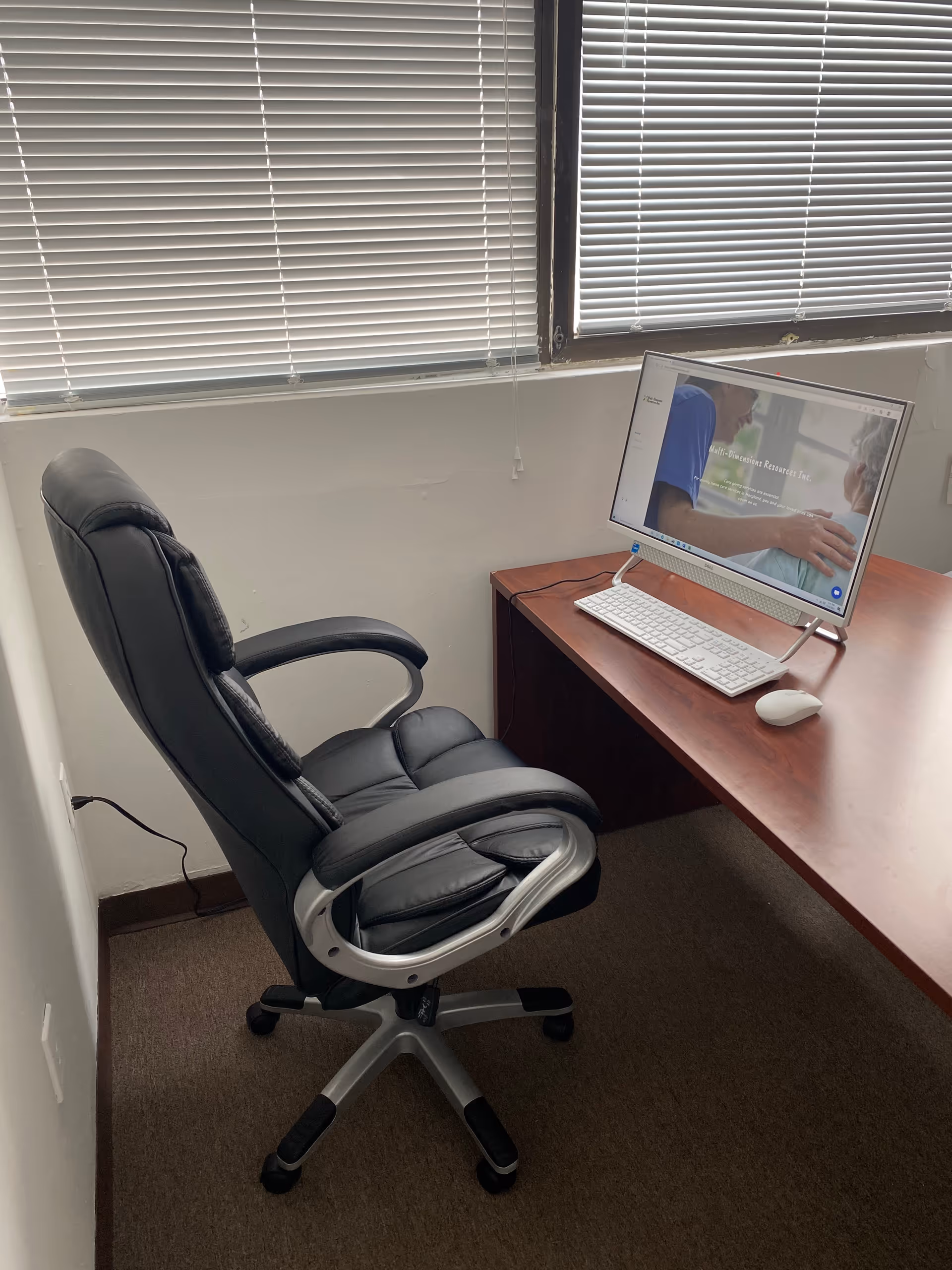Ergonomic black office chair at a wooden desk with a computer under blinds.
