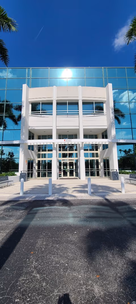 Modern glass-front building entrance with palm trees and a sunny blue sky.