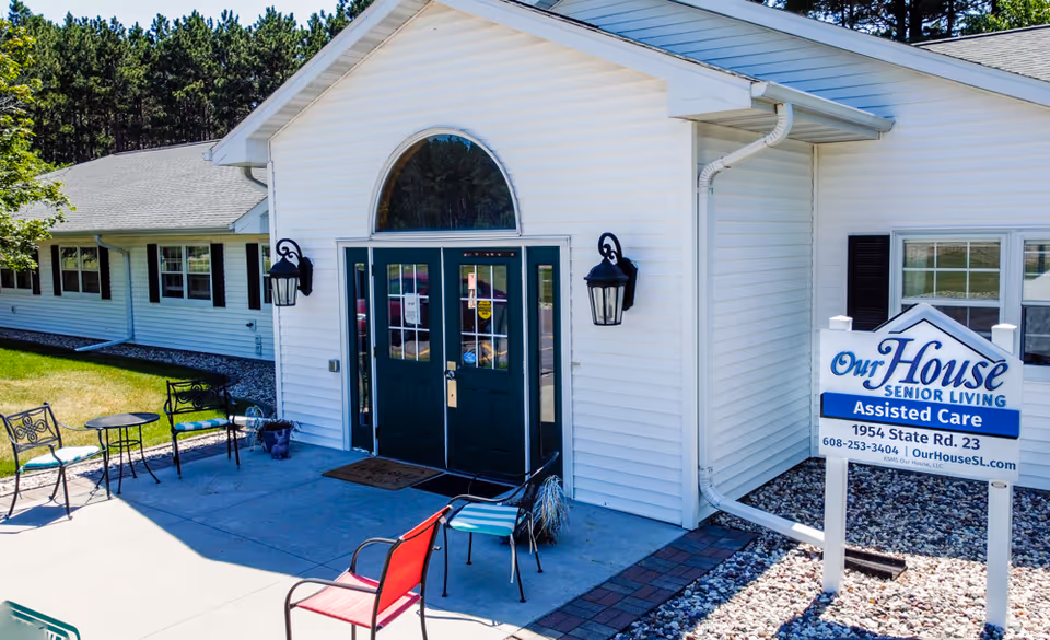 Exterior view of Our House Senior Living - Wisconsin Dells Assisted Care facility showing white building with dark green double doors, outdoor seating with metal chairs and tables, and a sign with the facility name, address, phone number, and website.