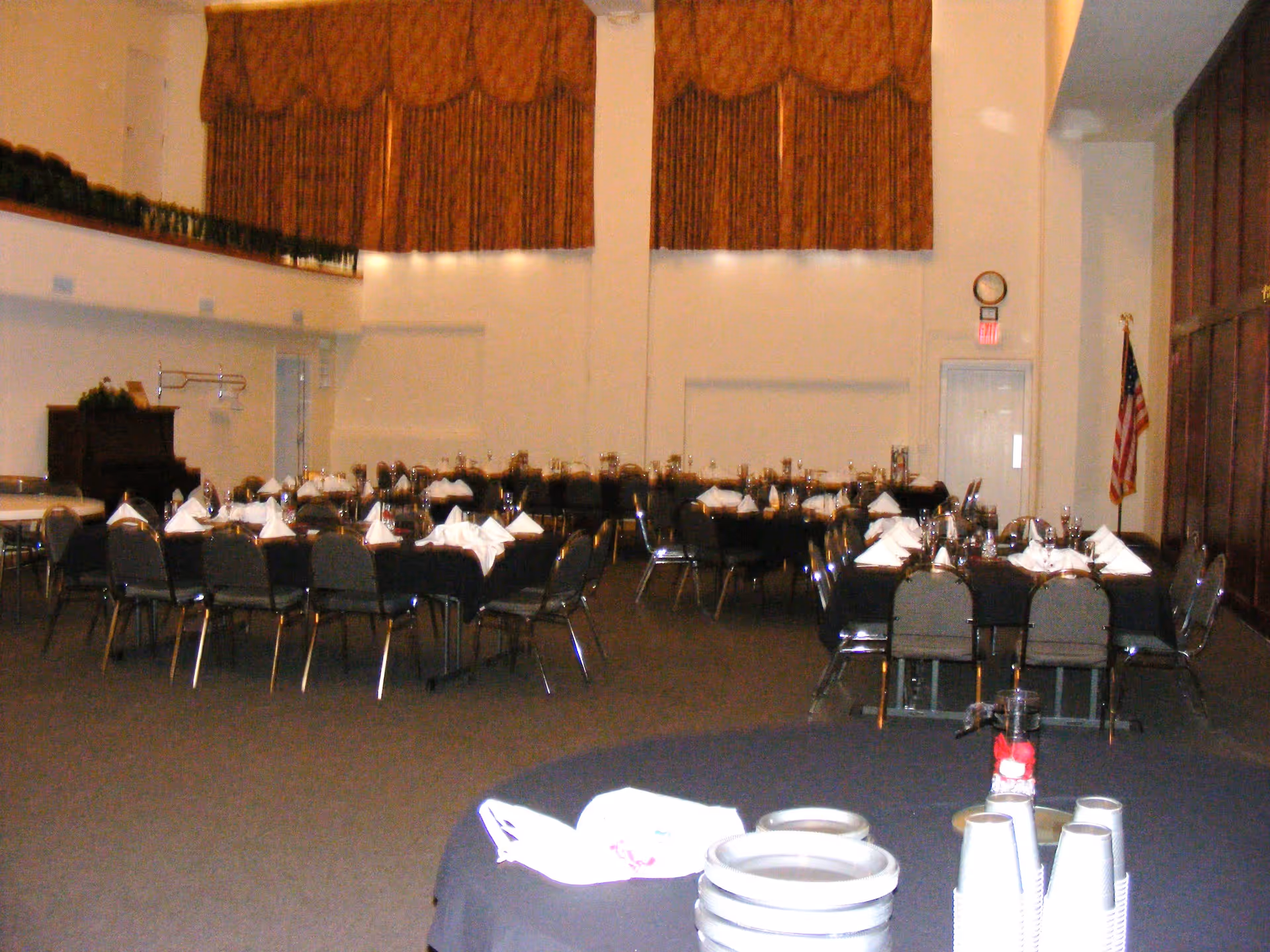 A large dining room set up for an event with multiple tables covered in black tablecloths, each with neatly folded white napkins and glassware. The room has high ceilings with large brown curtains on the far wall, an American flag near a door, and a piano on the left side. In the foreground, there is a round table with stacked plates and disposable cups.
