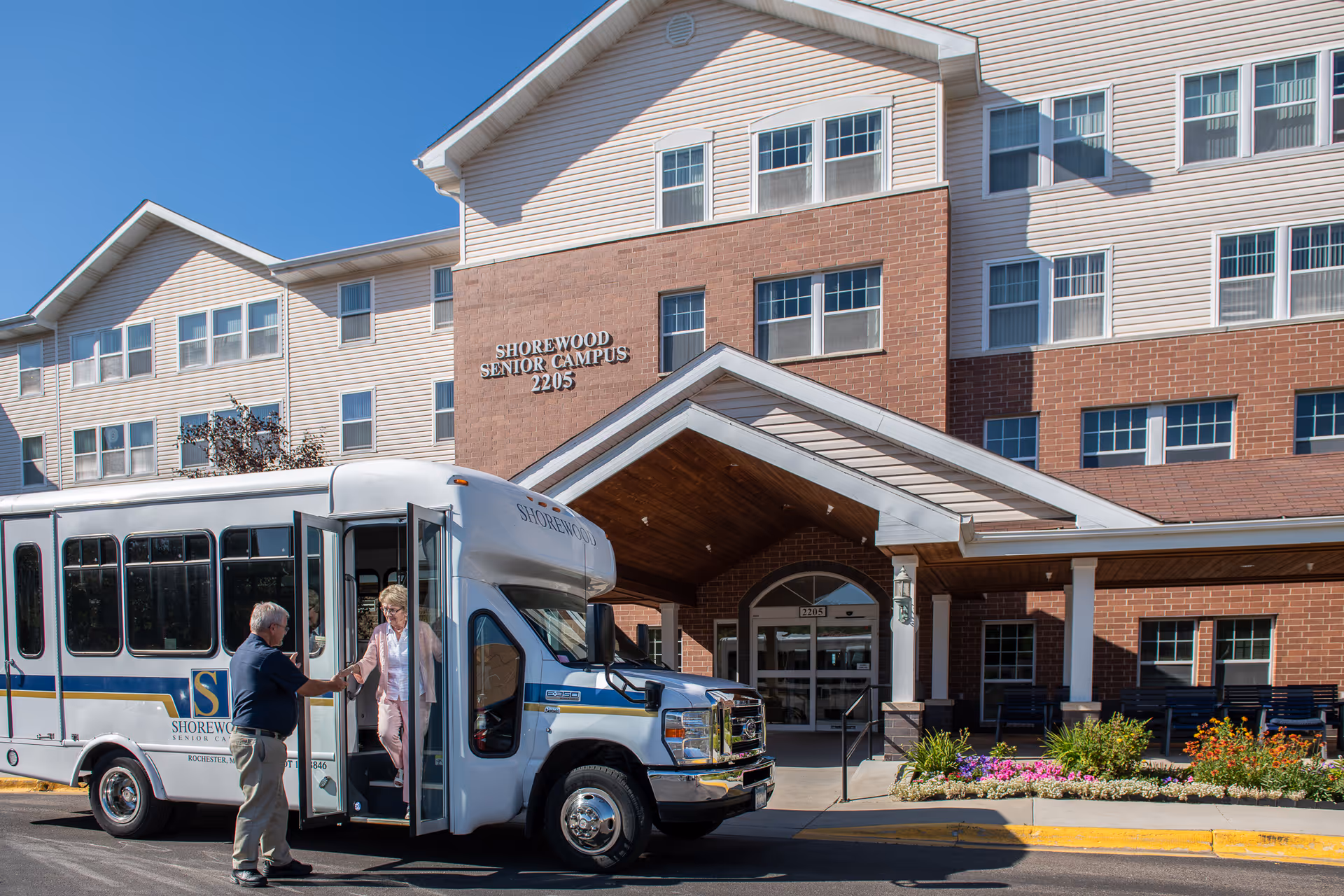 A woman is stepping off a white Shorewood Senior Campus shuttle bus and being assisted by a man outside the entrance of a multi-story senior living facility building with brick and beige siding, under a clear blue sky.