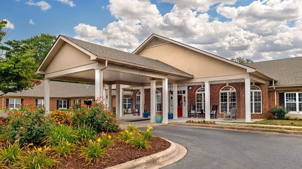 Exterior view of a senior living facility entrance with a covered drop-off area supported by white columns. The building features brick and beige siding with large windows and rocking chairs on the porch. There are landscaped flower beds with green shrubs and yellow flowers in front of the entrance under a partly cloudy sky.