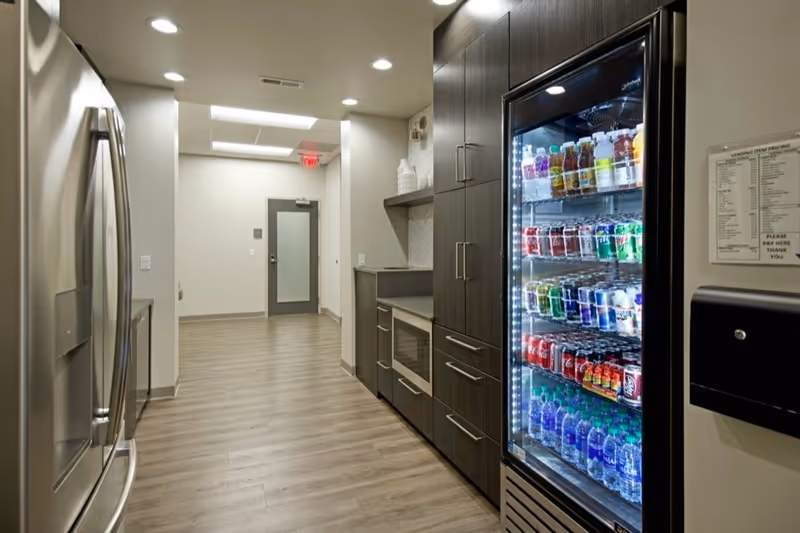 Modern staff kitchen with stainless refrigerator, beverage cooler, and dark cabinets along hallway