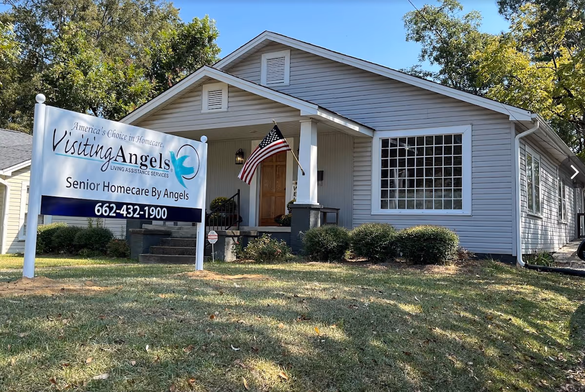 A single-story gray building with a front porch, sign, and American flag in a grassy yard.