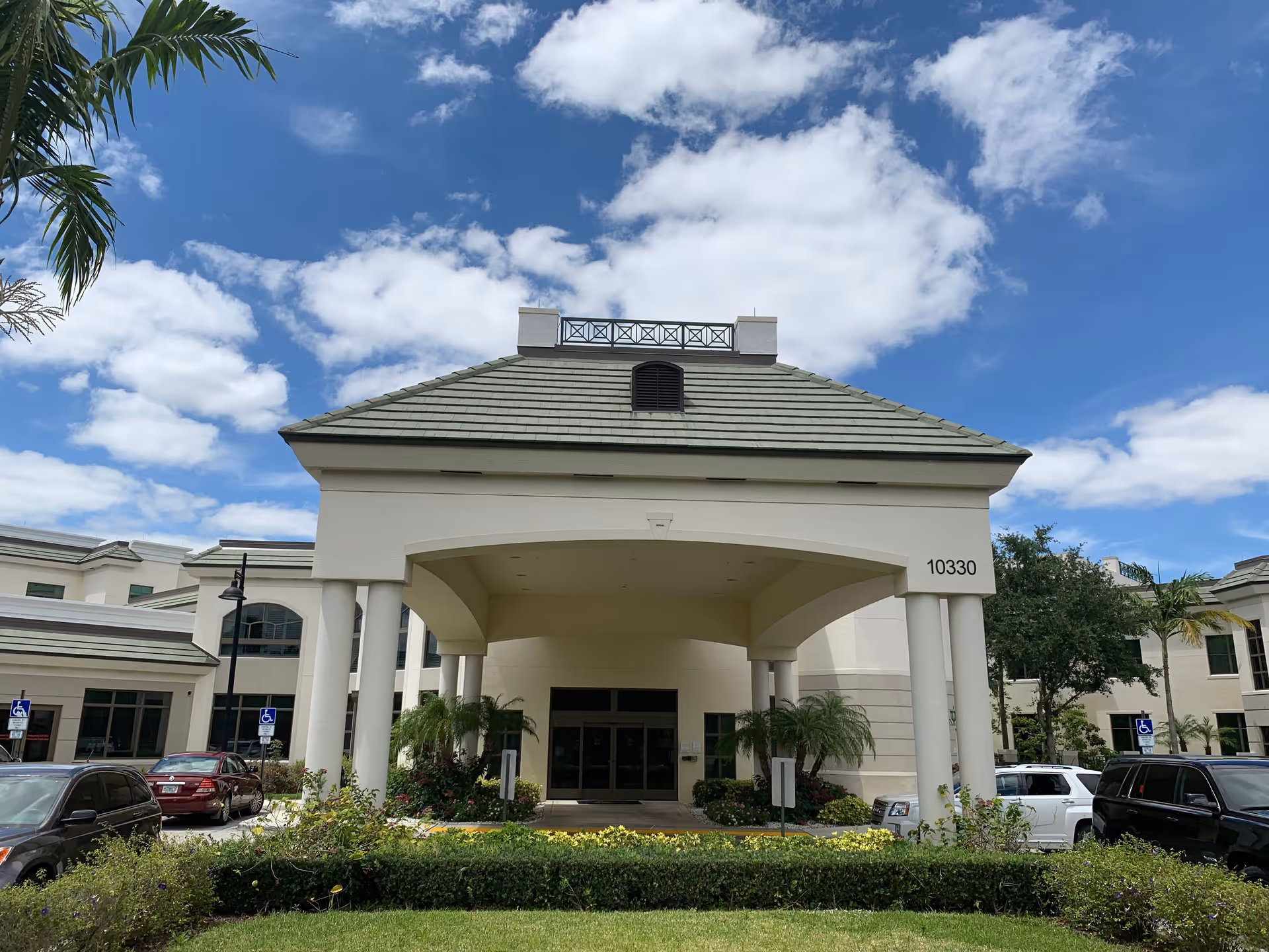 Front entrance of a light-colored senior living building with a covered porte-cochère, columns, address number 10330, parked cars and palm trees under a partly cloudy sky.