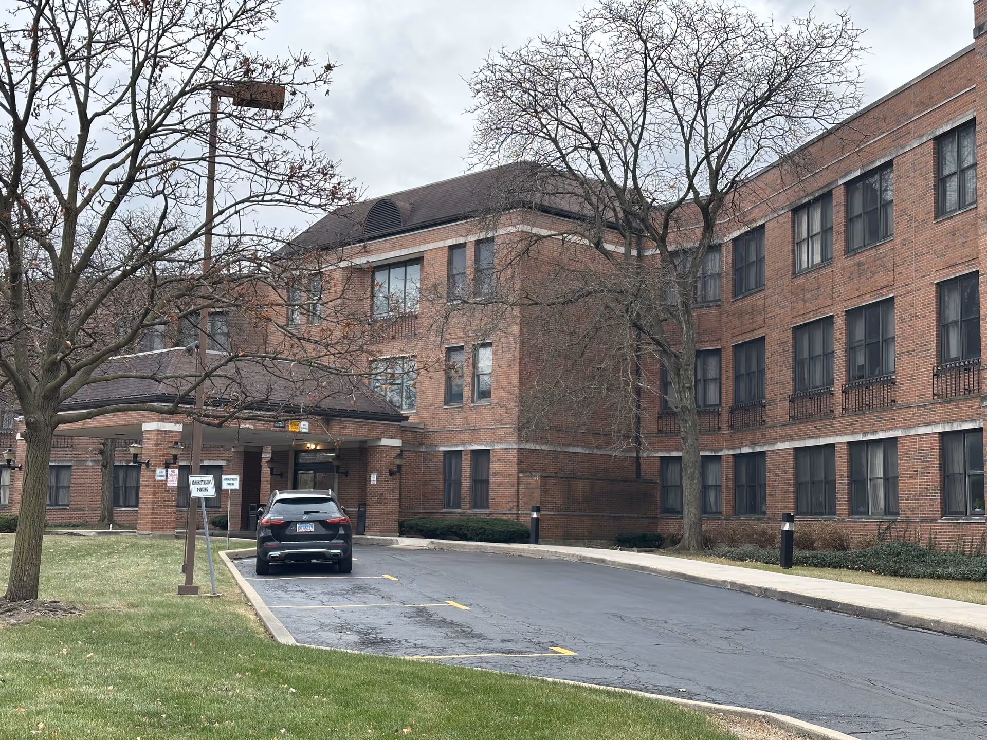 Exterior view of a three-story brick building with multiple windows, a covered entrance with a driveway, a parked black SUV, leafless trees, and a grassy area in front under a cloudy sky.