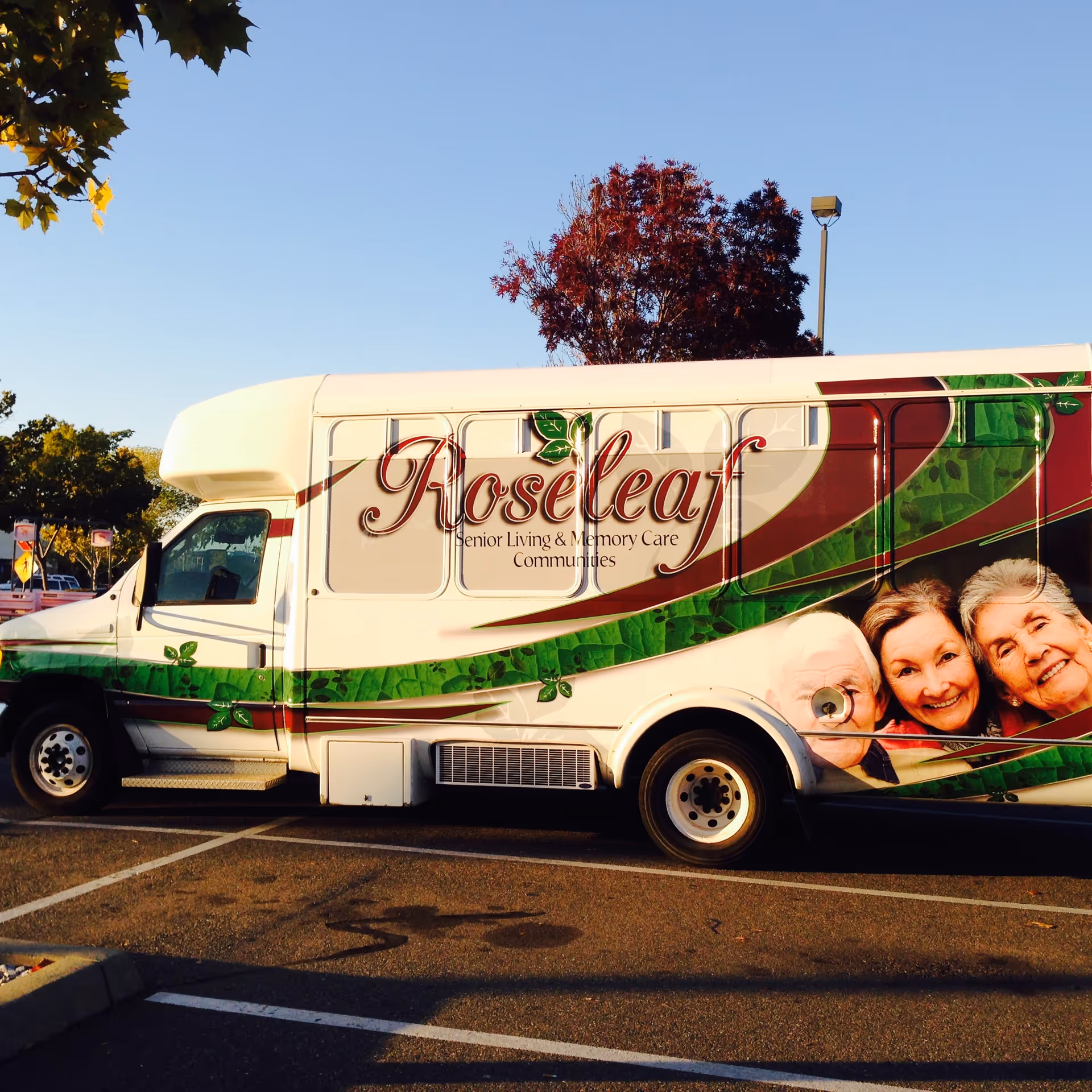 A white shuttle bus parked in a lot with the name 'Roseleaf Senior Living & Memory Care Communities' on the side. The bus features a green and maroon leaf design and images of three smiling elderly people on the rear side. Trees and a clear blue sky are visible in the background.