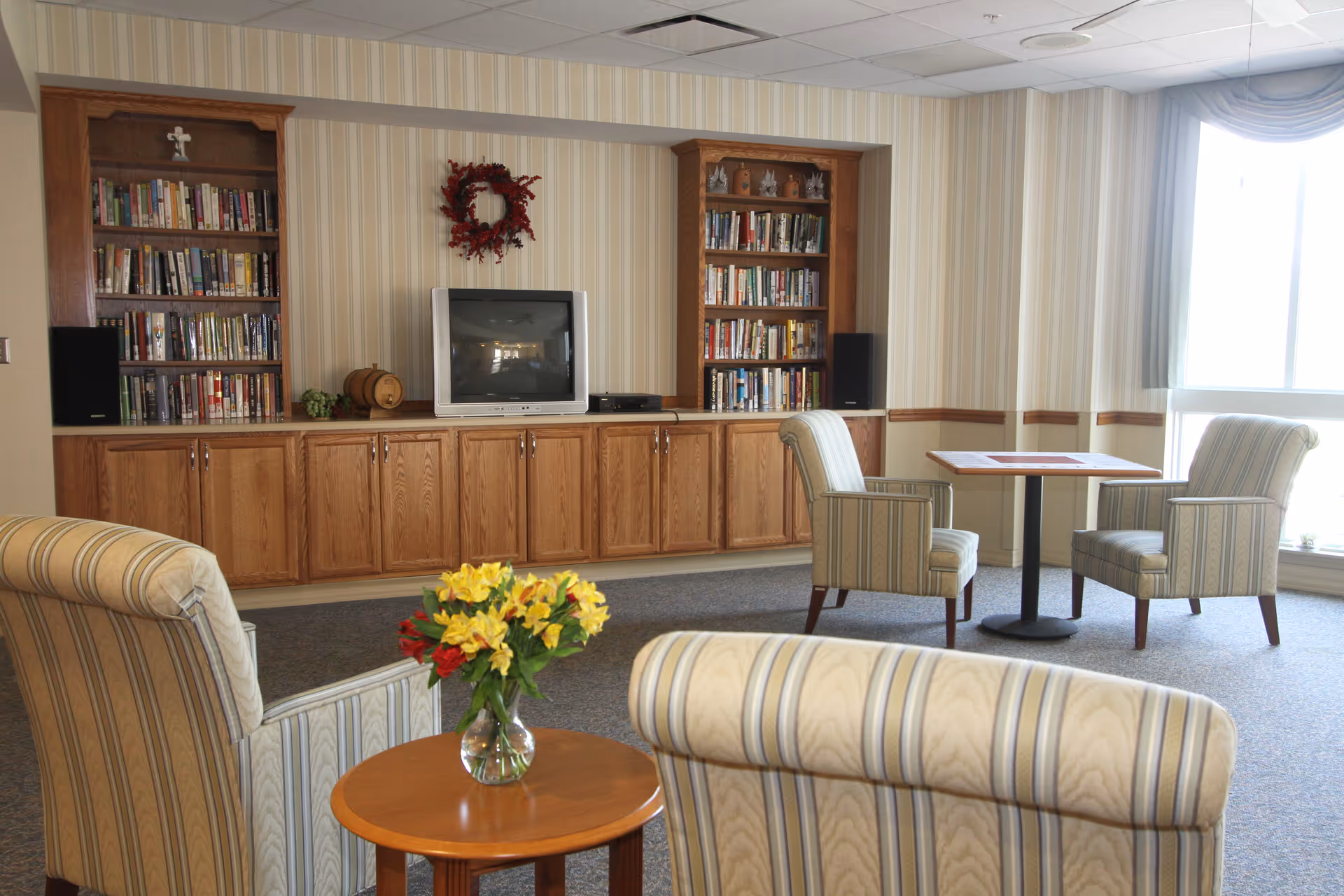 Cozy common living room with striped chairs, a TV, bookshelves, and a vase of flowers.